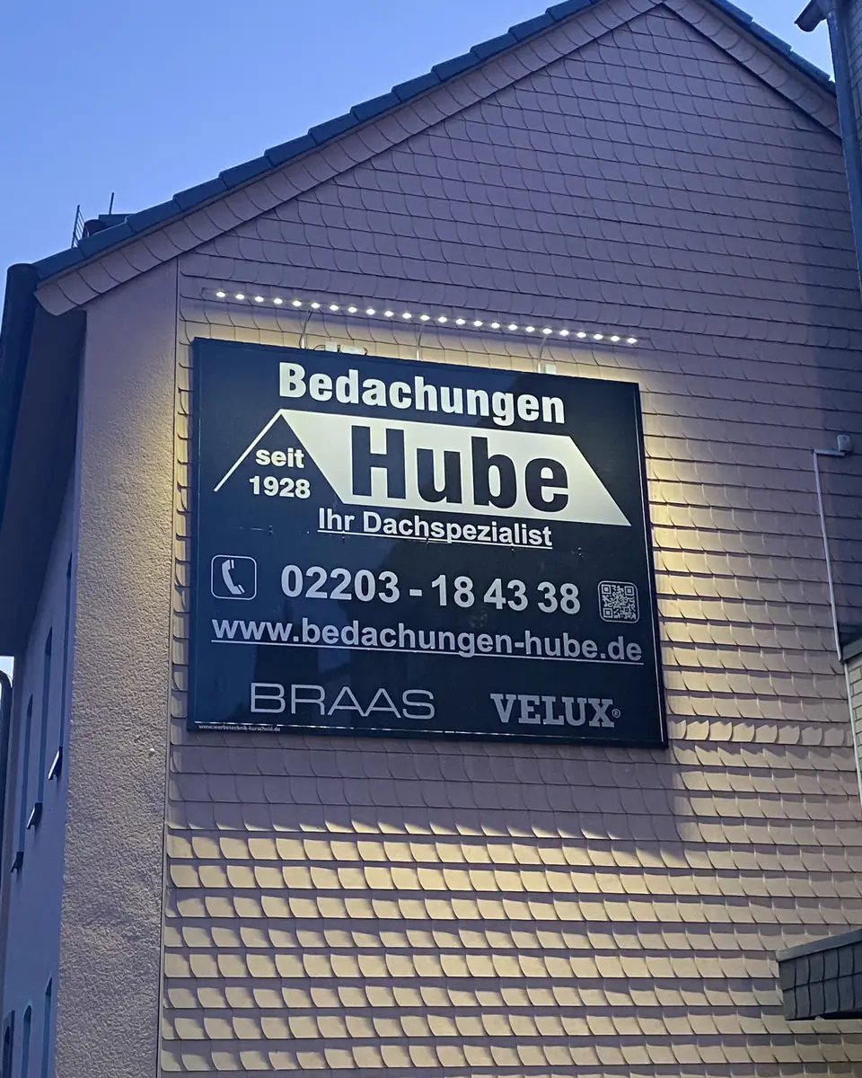 Das beleuchtete Schild von Bedachungen Hube an der Schieferfassade, abends